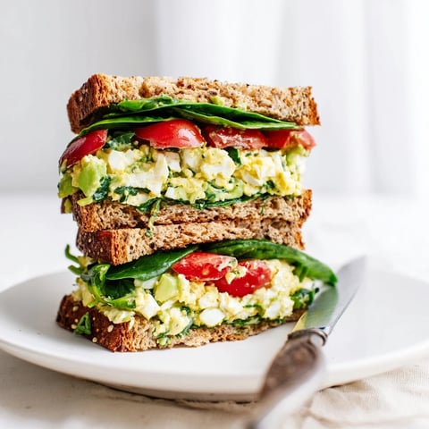 Avocado egg salad sandwich with creamy filling on toasted whole grain bread, topped with fresh spinach and ripe tomato slices.
